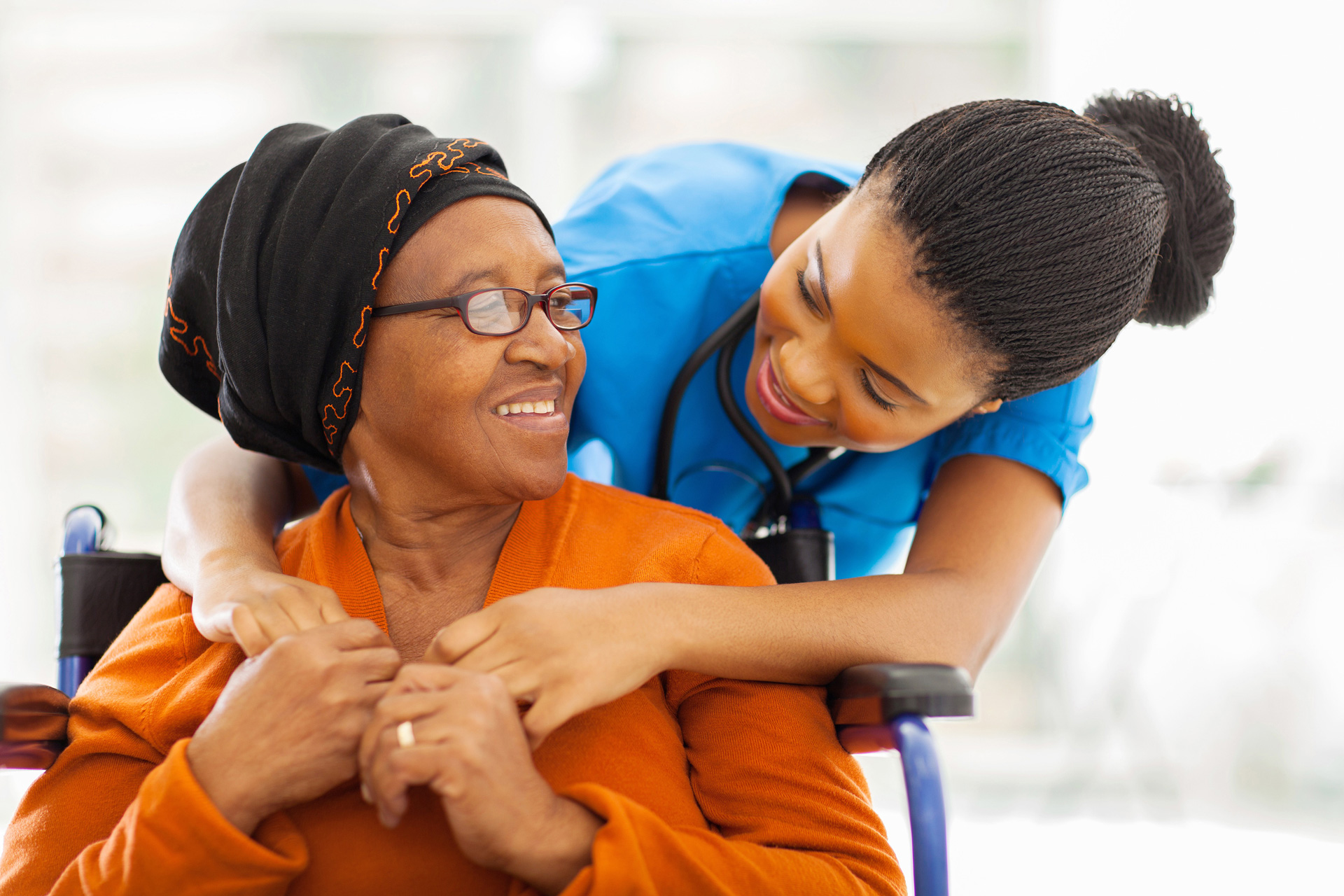 smiling nurse in blue scrubs leaning to smiling woman in wheelchair